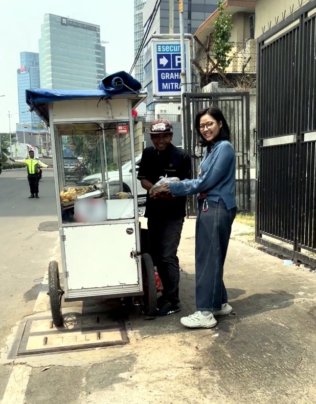 Relawan menyerahkan nasi kotak kepada pedagang gerobak di trotoar, dengan gedung perkantoran di latar belakang