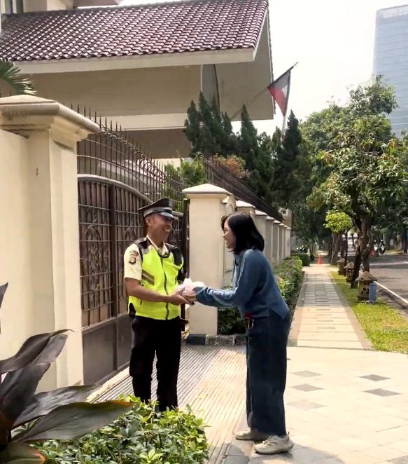 Petugas keamanan berompi kuning tersenyum menerima nasi kotak dari relawan di depan pagar rumah
